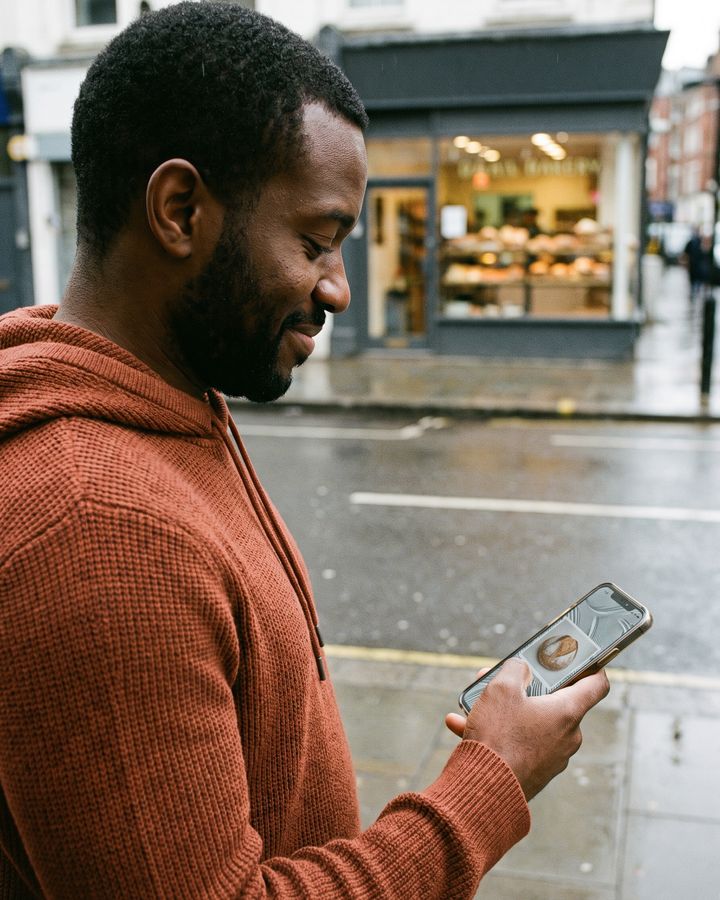 A young man on the sidewalk nods at a clear answer on his phone, with the bakery across the street